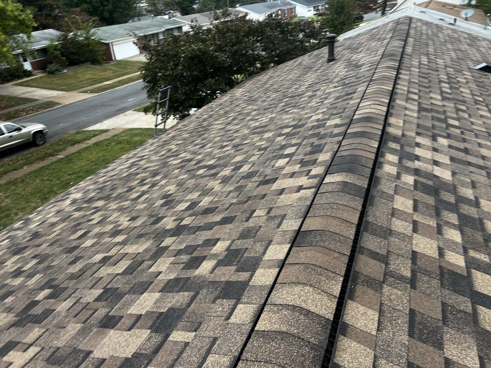 Brown, tan and charcoal multi tone architectural shingles running to a capped ridge with a dark ridge vent slot beneath the cap, a metal flue cap near the far end, and an aluminium ladder standing against the eave at the upper left, above a suburban street of single storey brick and vinyl ranch houses