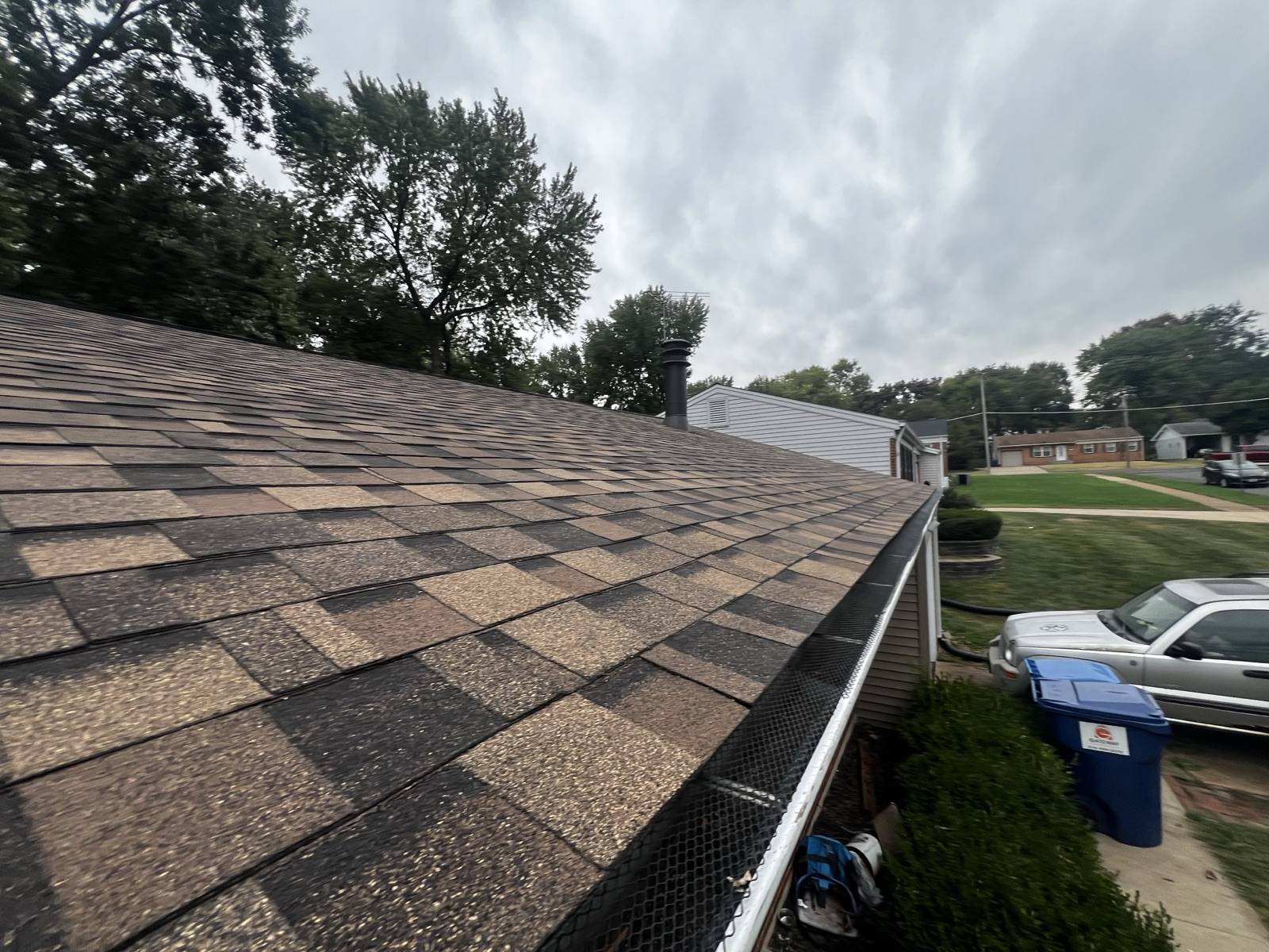 Worn brown and tan shingles seen low along the eave from the roof surface, granule loss showing as pale patches across the field, a black mesh gutter guard fitted along the white gutter, a metal flue cap at the ridge and a suburban ranch street with parked vehicles below